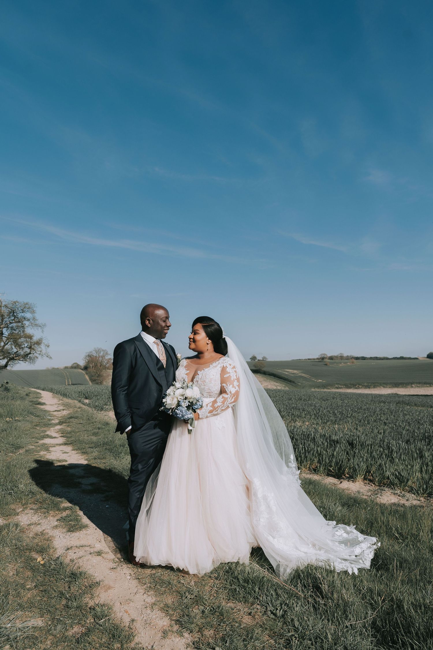 A wedding couple poses together in a scenic field with rolling hills and blue sky in the background.