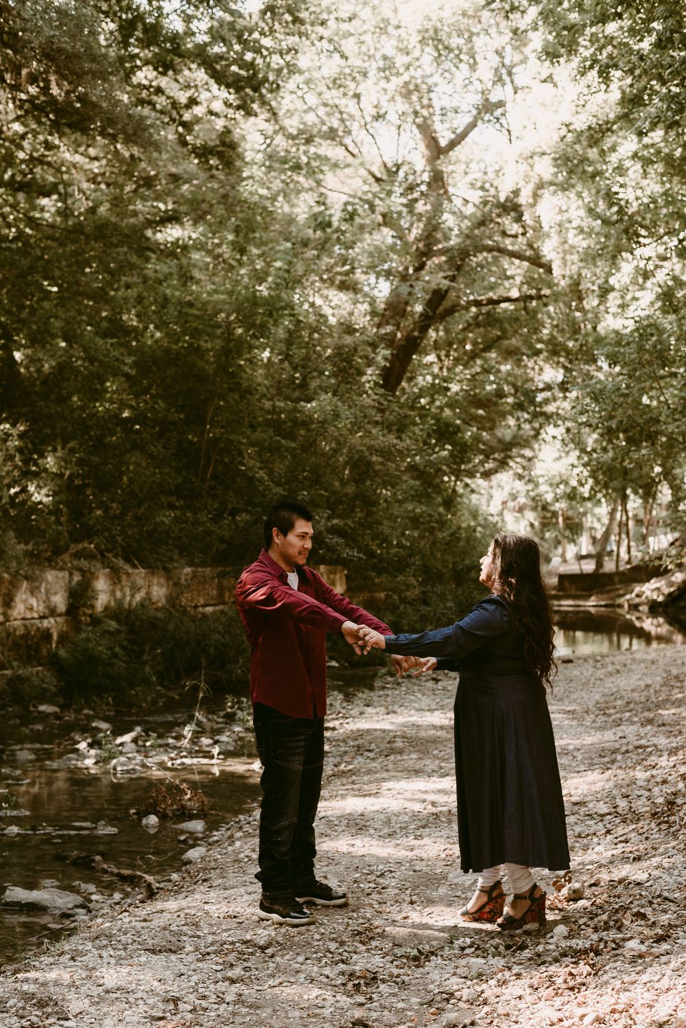 A couple holds hands while dancing on a nature trail next to a peaceful stream.