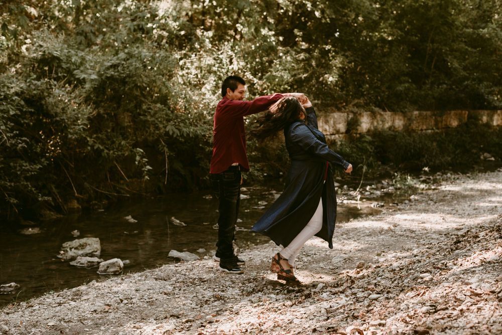 A couple shares a playful dance moment by a stream in a wooded setting.