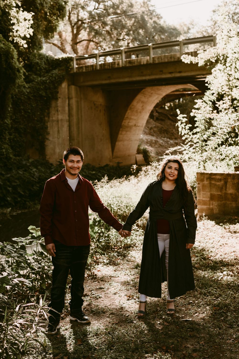 A couple holds hands while standing beneath a large stone bridge in a lush outdoor setting.
