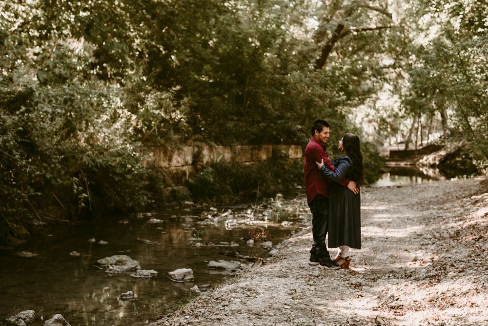 A romantic couple embraces on a wooded path beside a tranquil stream in moody autumn lighting.