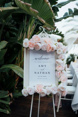 Elegant wedding welcome sign decorated with blush and white roses against tropical greenery.