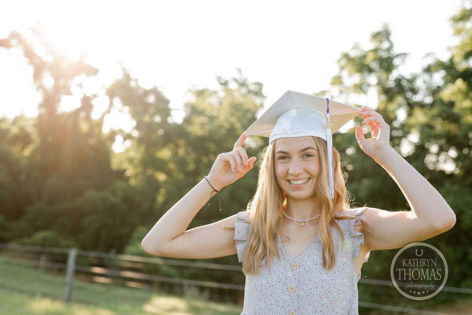 A graduate playfully adjusts their cap while standing in a field at sunset.