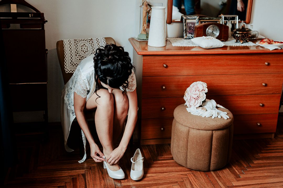 A white wedding shoe being placed on a foot next to a wooden dresser.