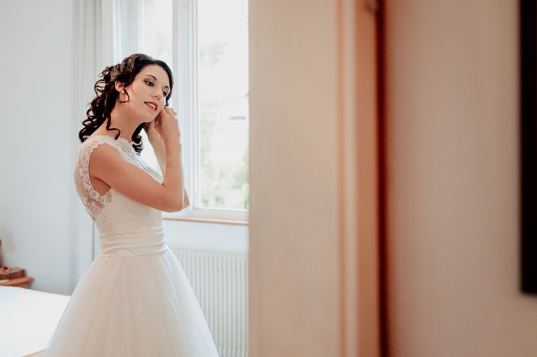 Bride in white wedding dress looking pensively out window during preparation.