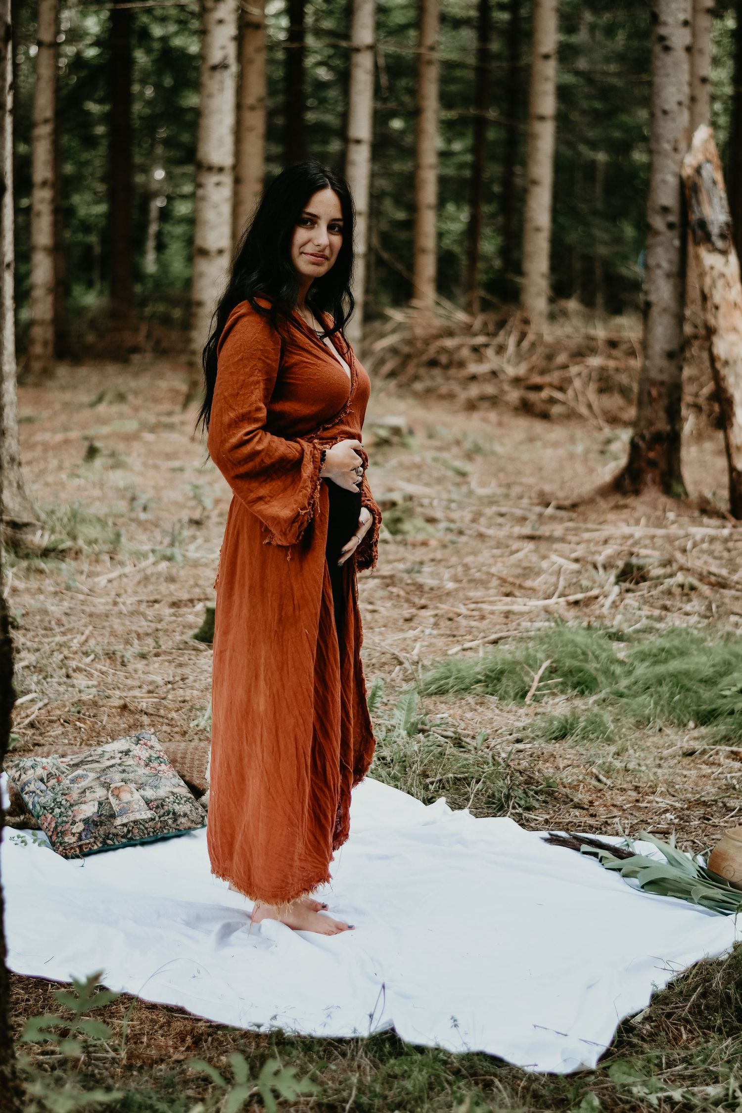 A figure in a flowing rust-colored dress poses in a misty forest setting on a white blanket.