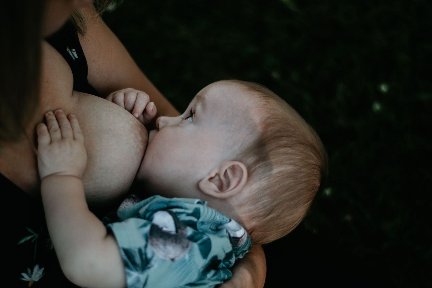 Close-up intimate nursing moment captured in low light with baby in floral outfit.