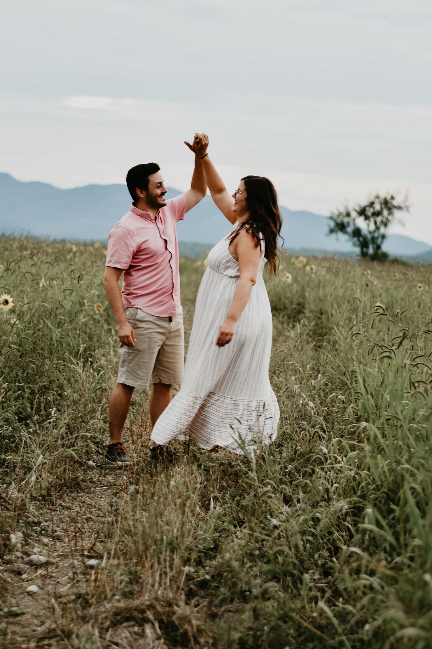 A couple dances joyfully in a mountain meadow at sunset, holding hands raised in celebration.