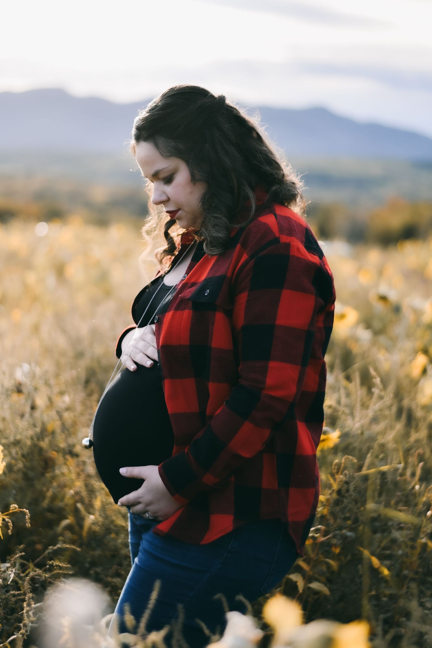 A person in a red and black buffalo plaid jacket stands in a golden field with mountains in the background.
