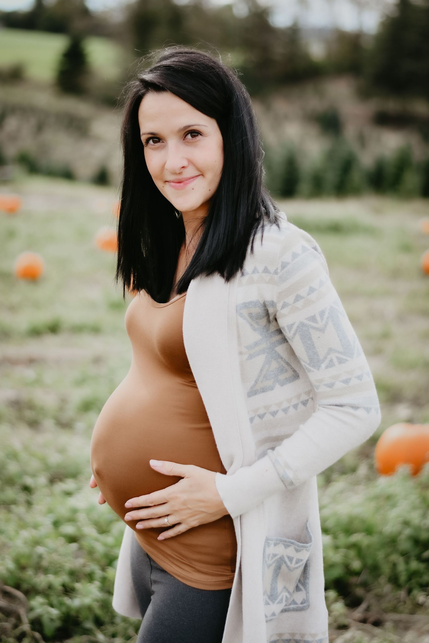An expectant pose in a pumpkin patch wearing a tan top and patterned cardigan.