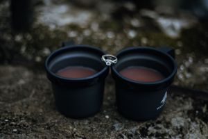 Two black camping mugs filled with hot drinks resting on a rocky surface outdoors.