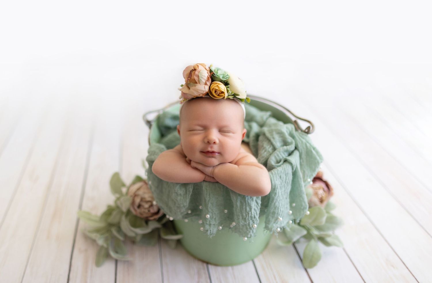 Sleeping newborn posed in mint green bowl with floral accents on white wooden floor.