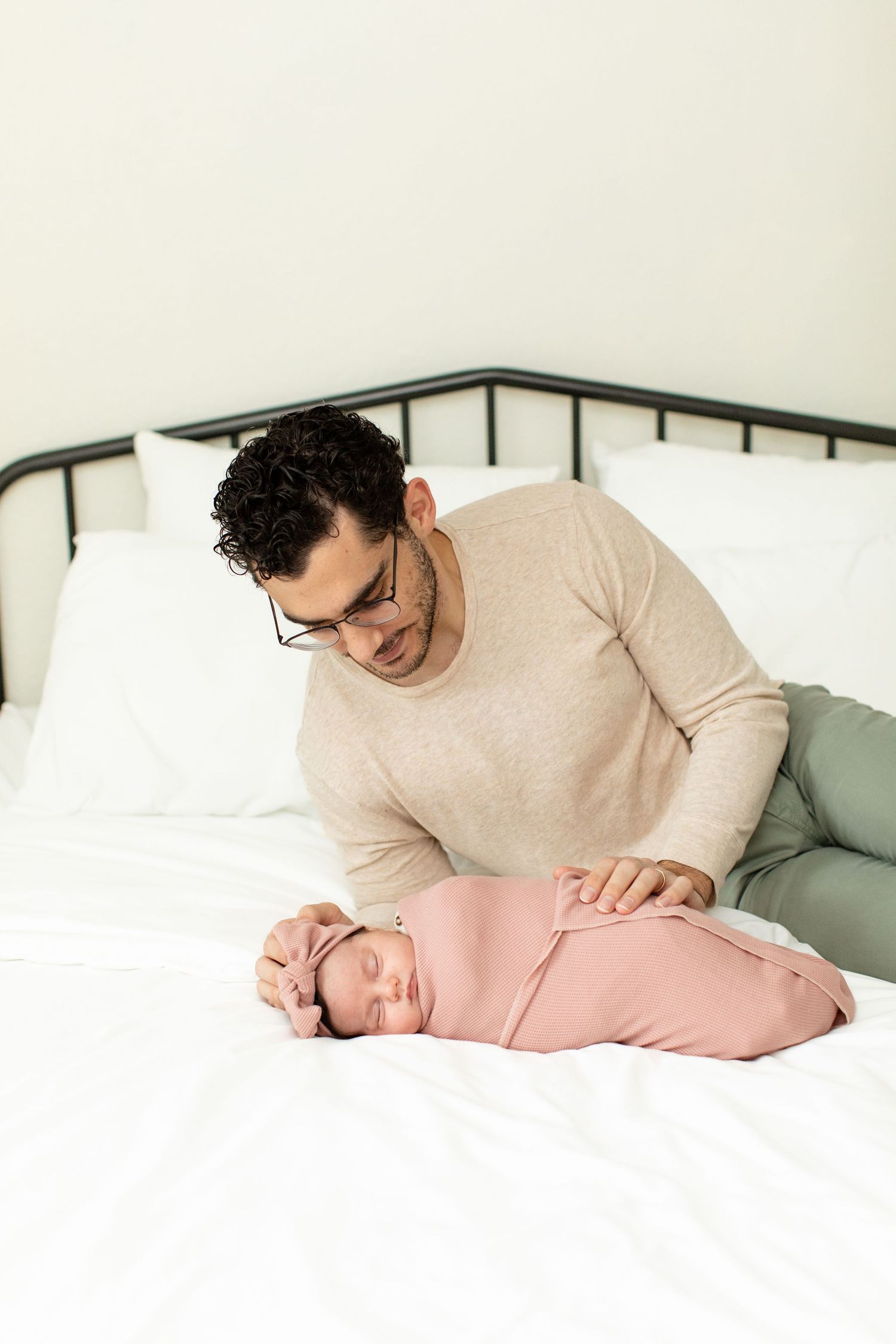 Man in beige sweater looking down at sleeping baby swaddled in pink on white bed.