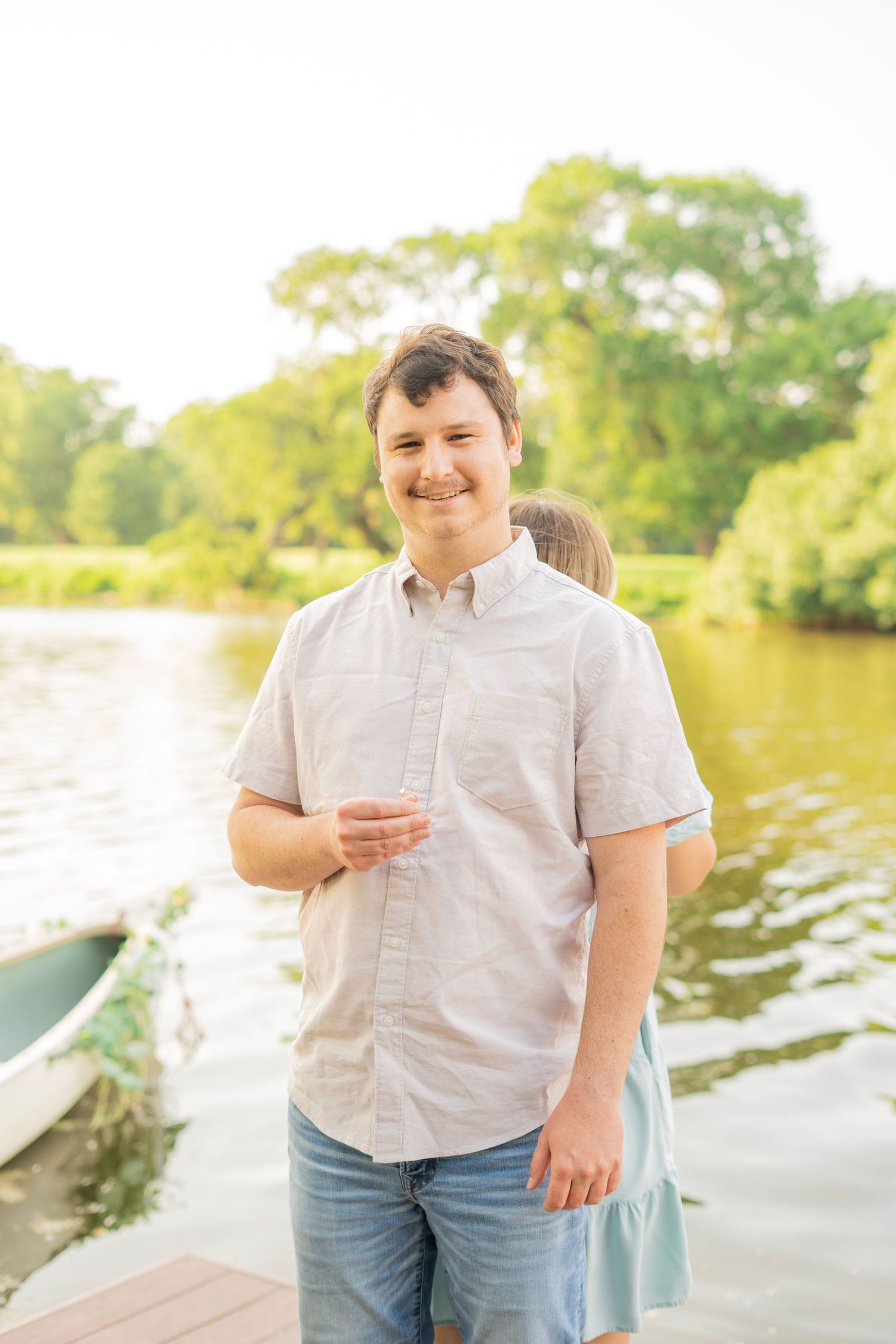 Smiling young man in a light shirt standing on a sunny dock beside a calm river.