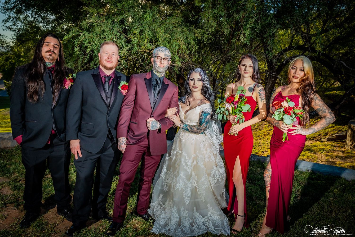 Wedding party posing outdoors in burgundy and white attire against green foliage.