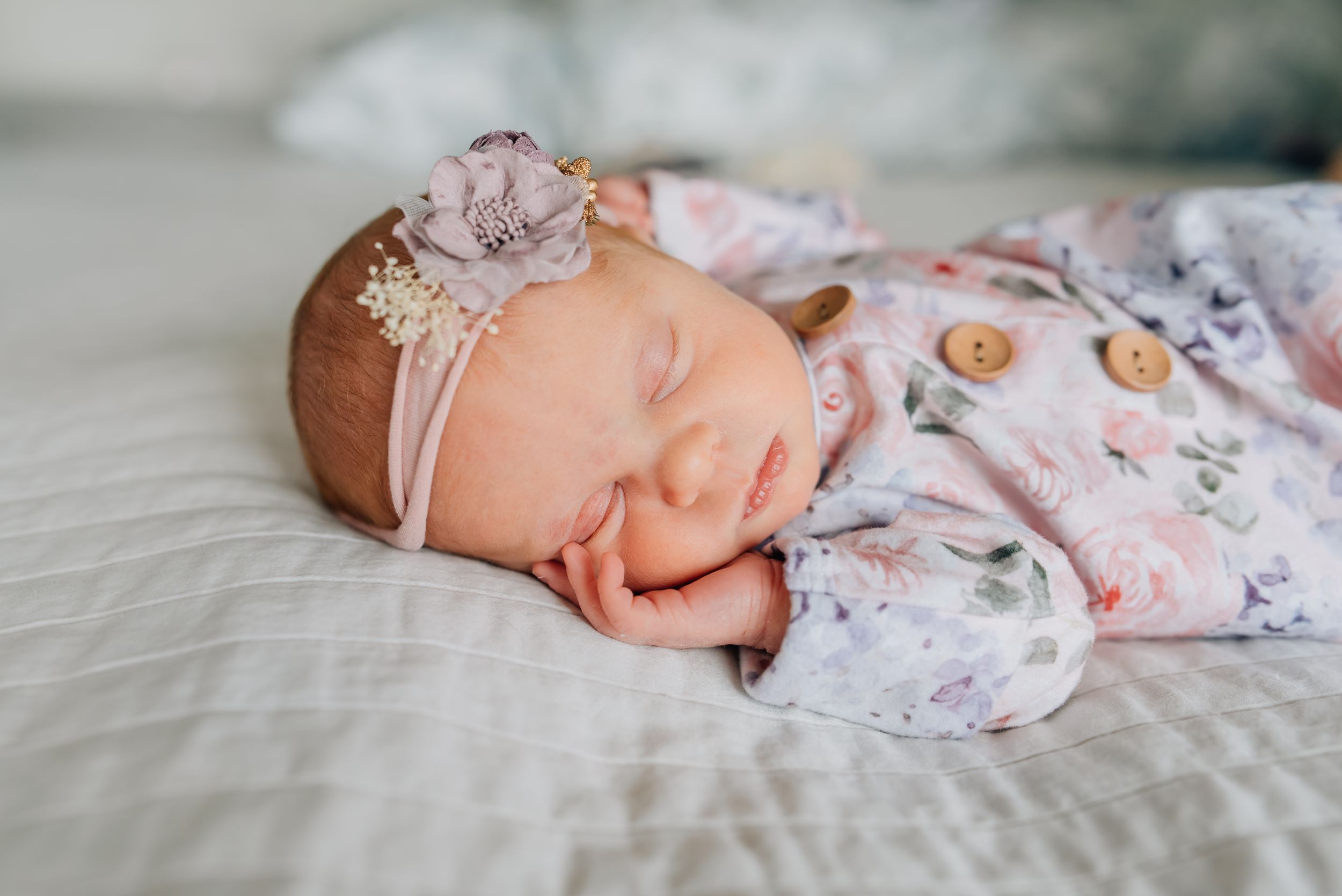 Newborn baby sleeping on her parents bed with a floral knotted gown and head band.