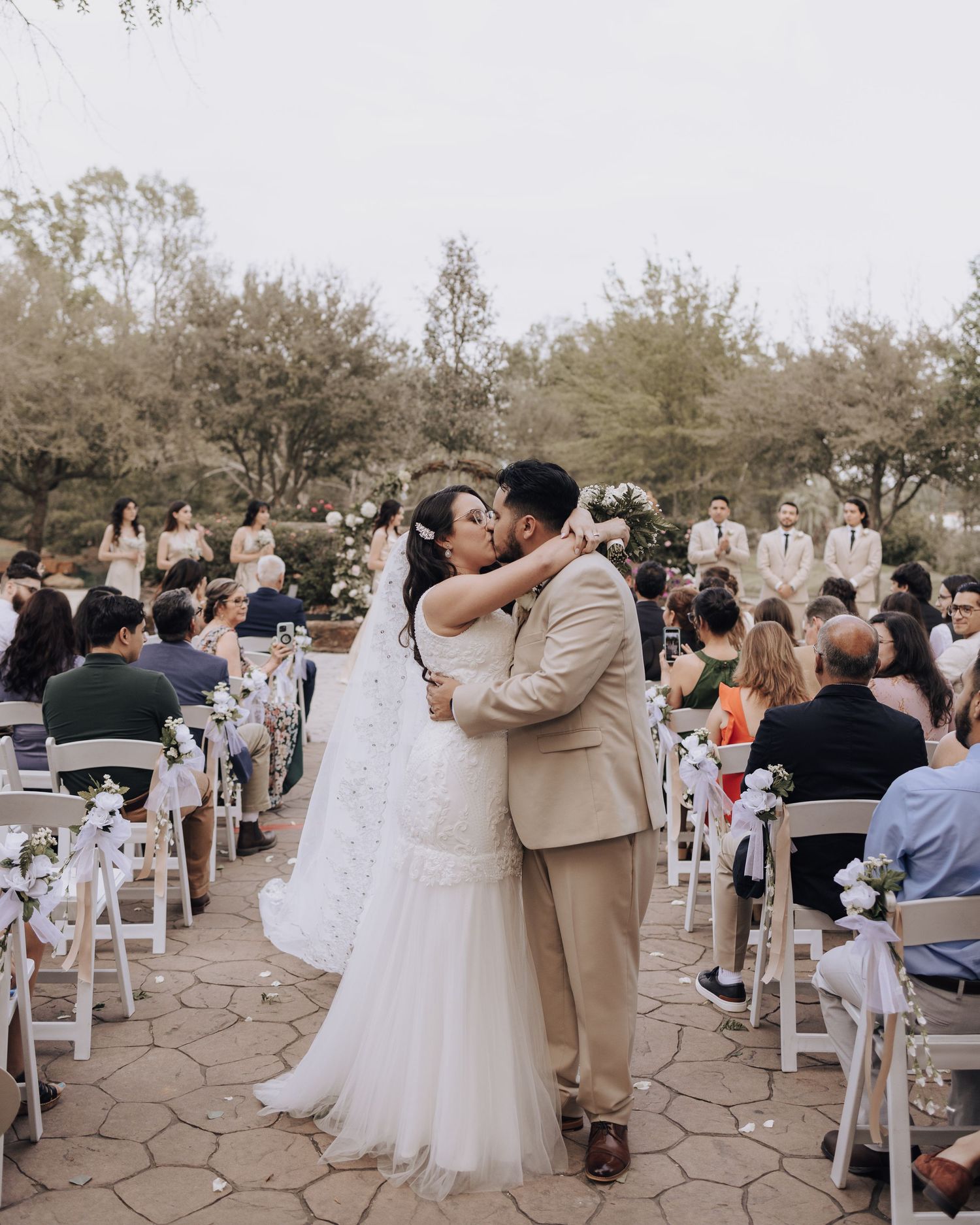 A romantic first kiss as newlyweds surrounded by guests in white chairs at an outdoor venue.