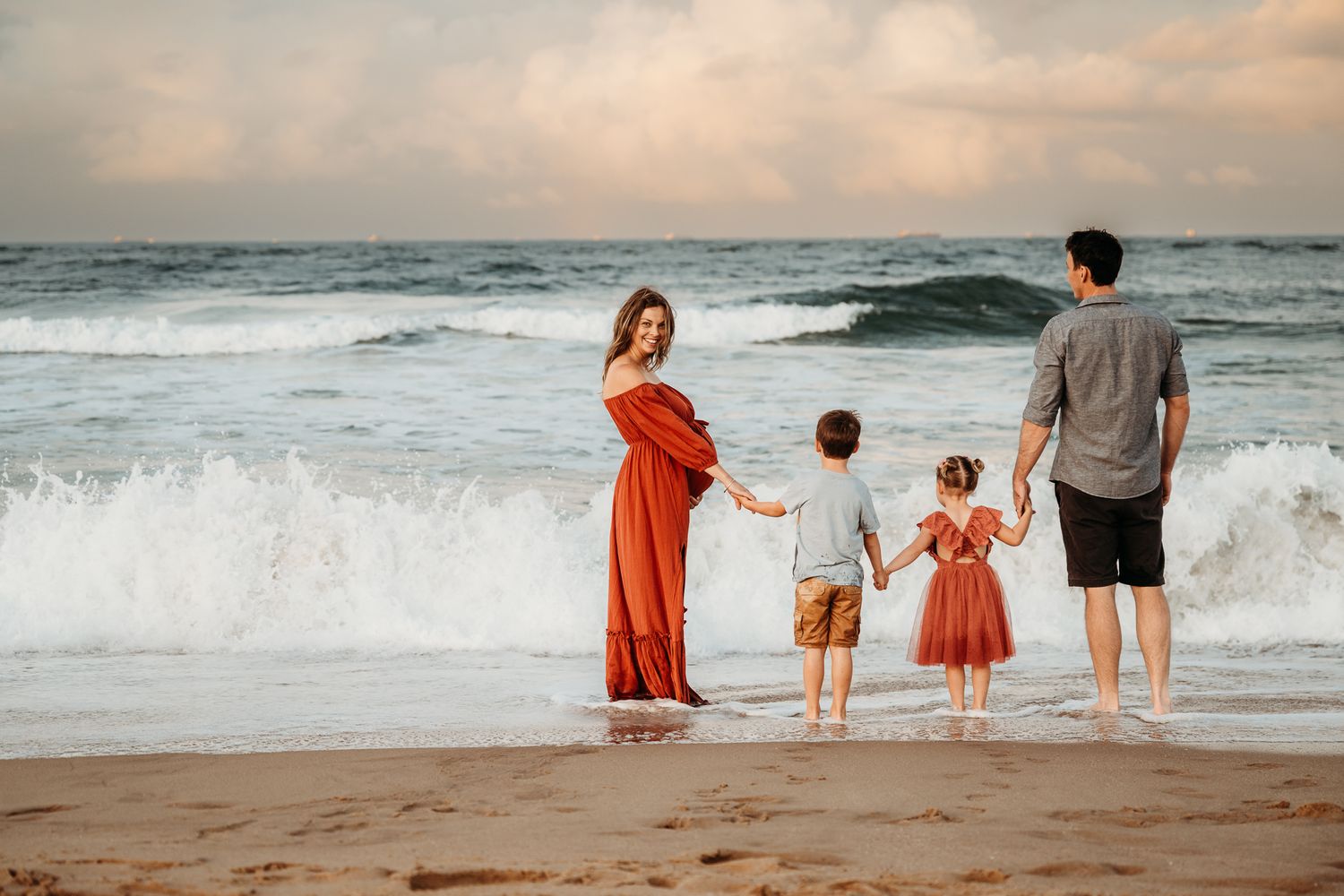 A family of four stands together on a beach at sunset as waves crash behind them.