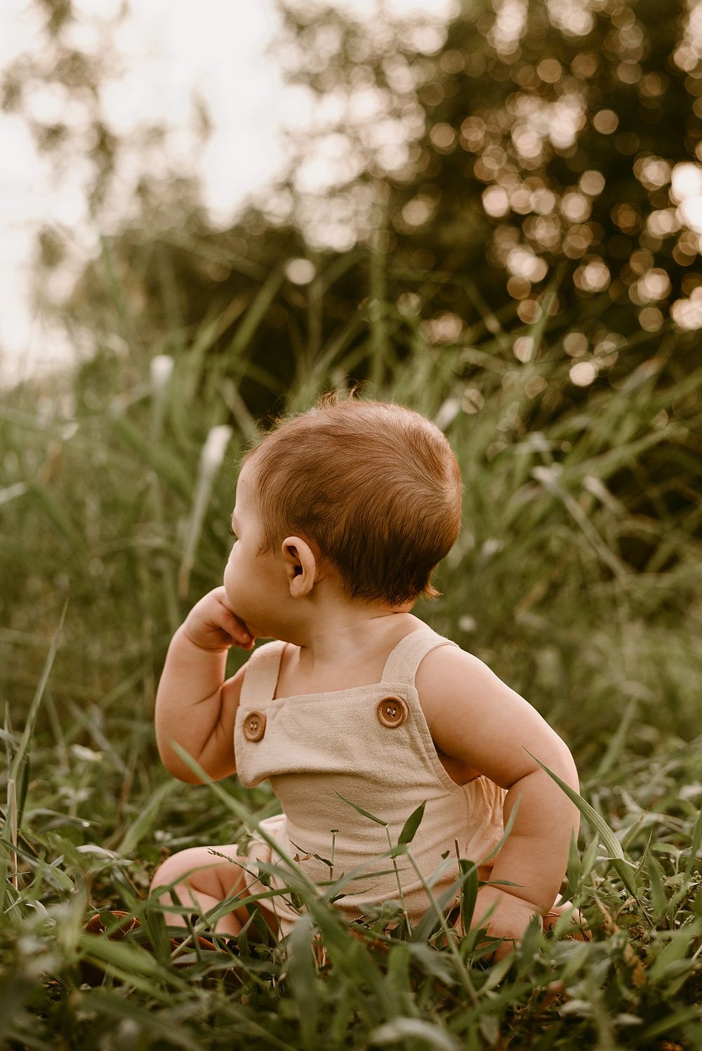 Baby in beige overalls touching face while sitting in grass with bokeh background.