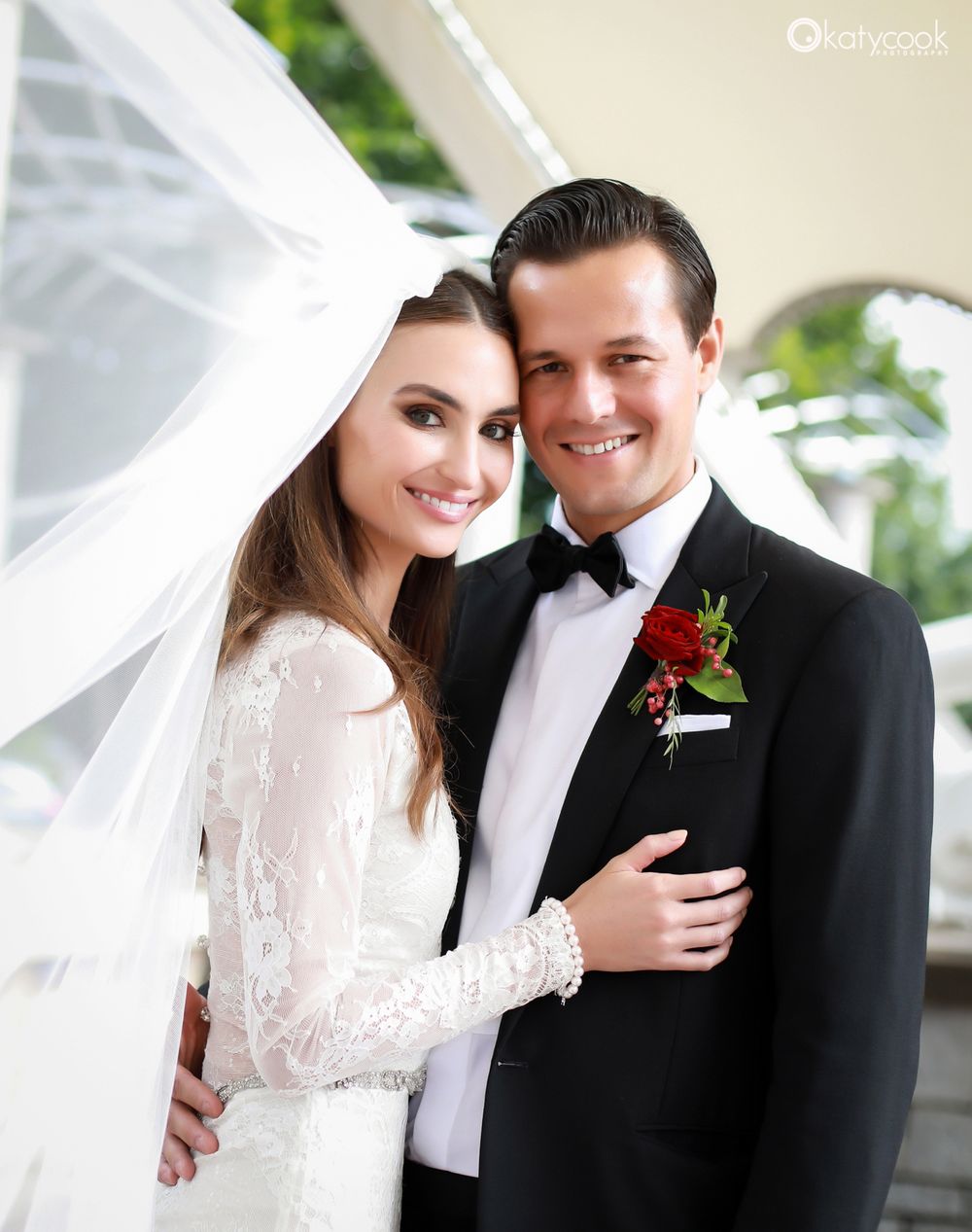 An elegant wedding photo featuring lace sleeves and a red boutonniere against a bright backdrop.