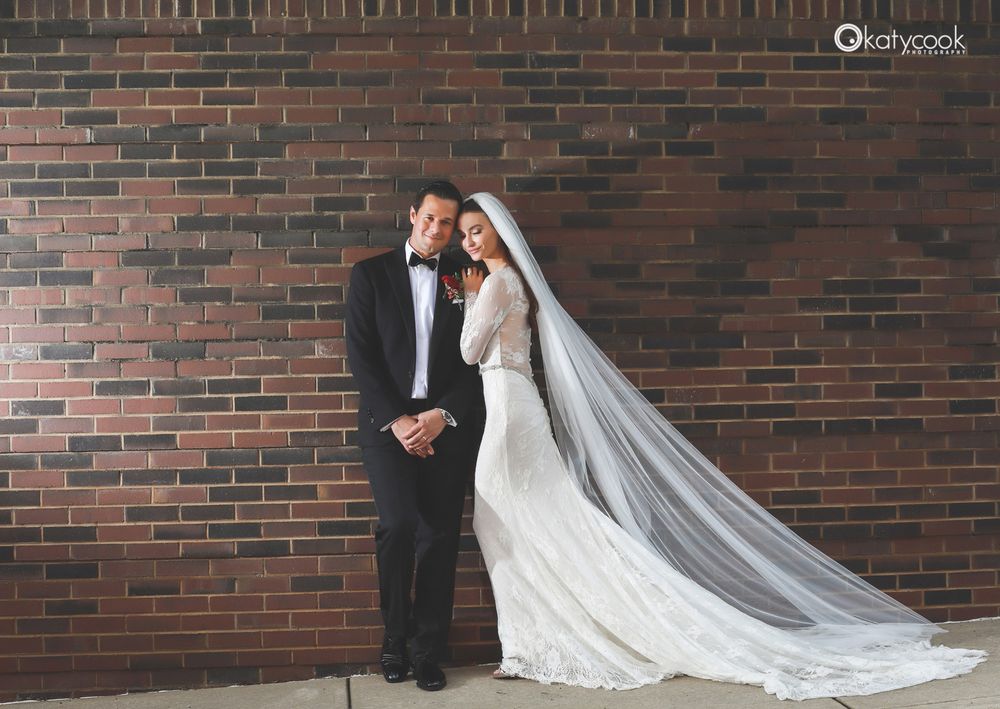 Newlyweds pose against a brick wall with a dramatic flowing cathedral veil.