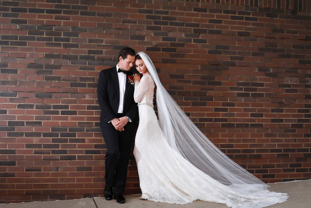 Wedding couple in formal attire standing against rustic brick wall backdrop.