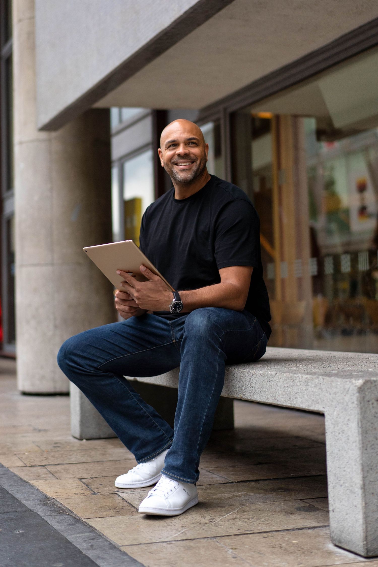 Business professional holding tablet while sitting on concrete ledge outside office building.