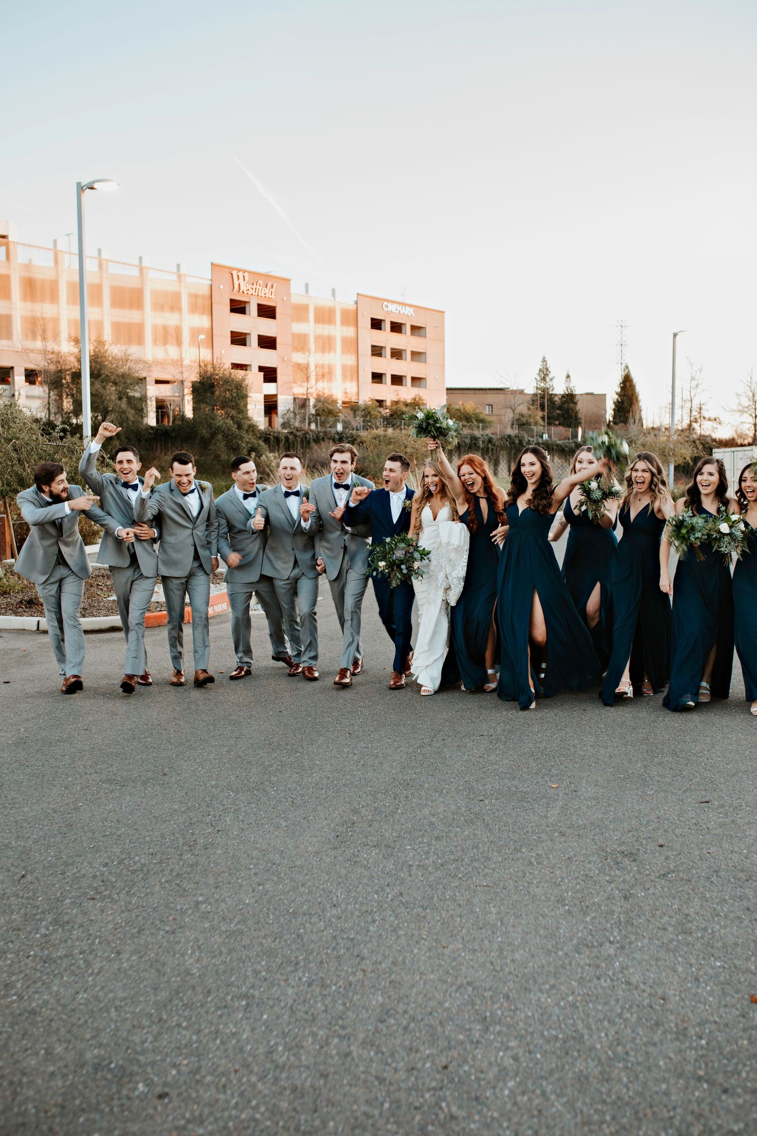 A wedding party poses together outdoors with bridesmaids in navy dresses and groomsmen in light gray suits.