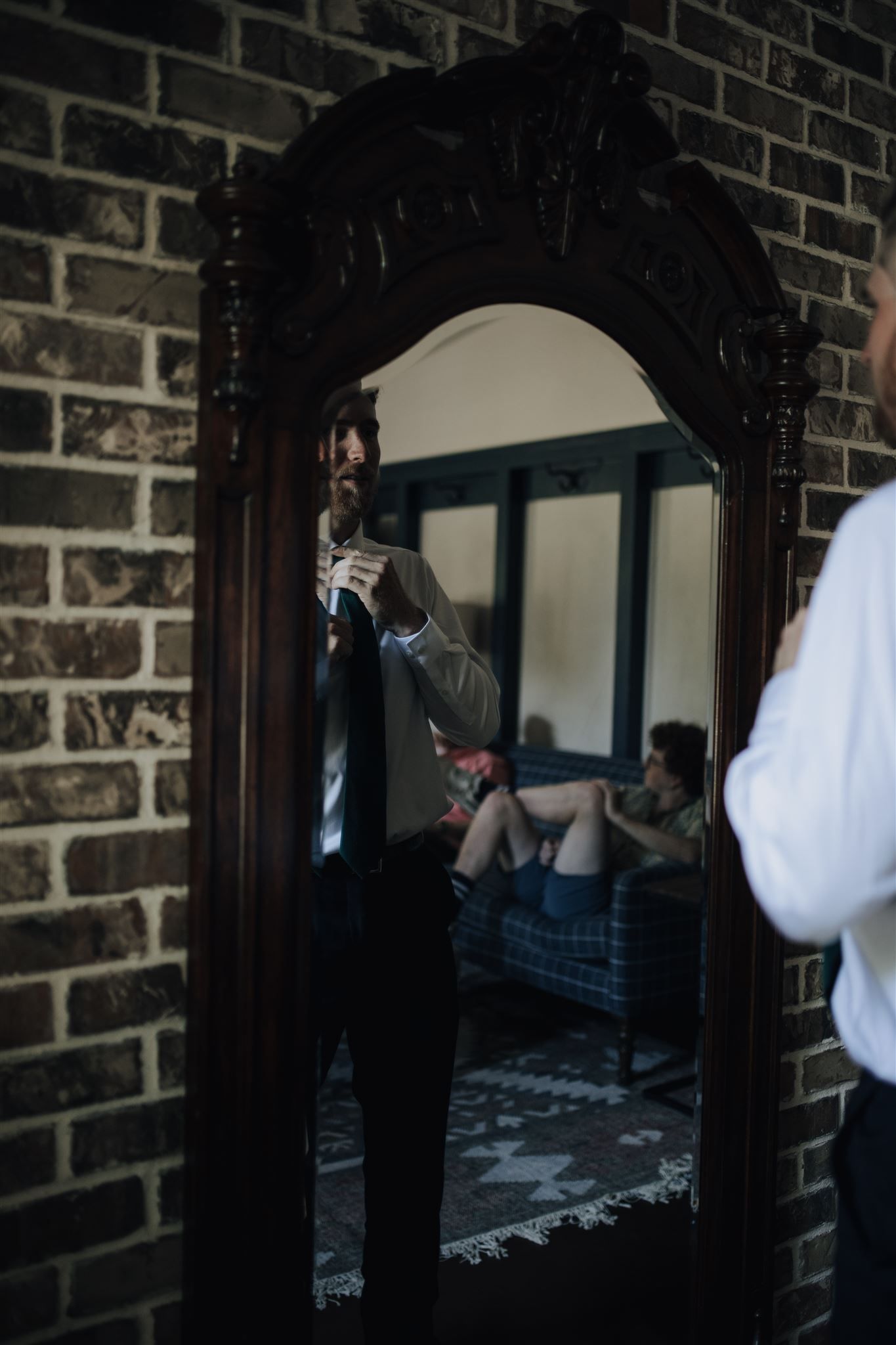 Man adjusts his tie in an ornate mirror reflection against exposed brick wall.