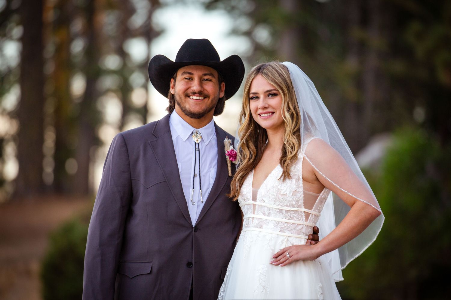 A newlywed couple poses in a forest setting, with the groom in a black cowboy hat and suit and the bride in a white gown and veil.