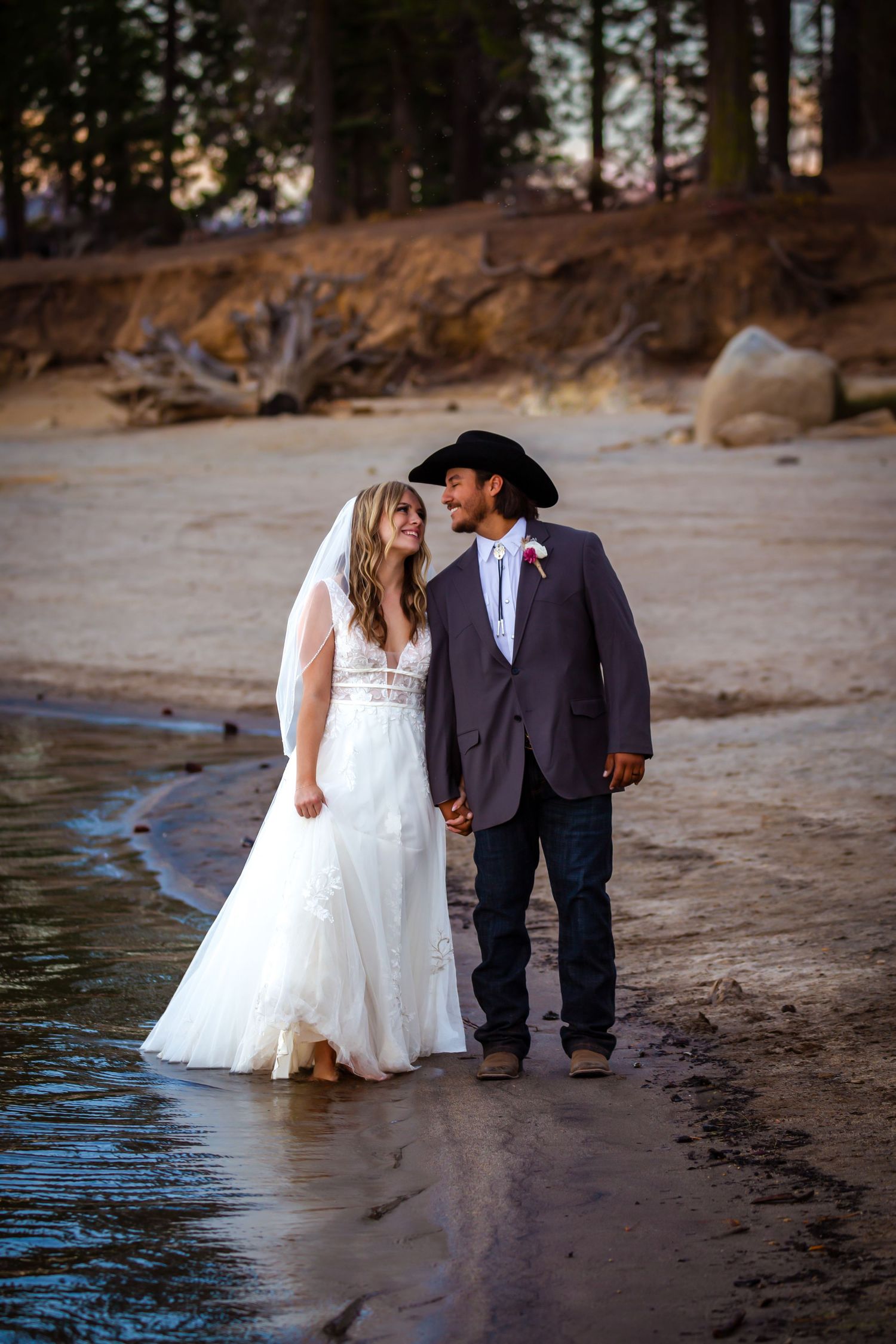 A couple in wedding attire walks along a sandy beach at sunset with water visible in the background.