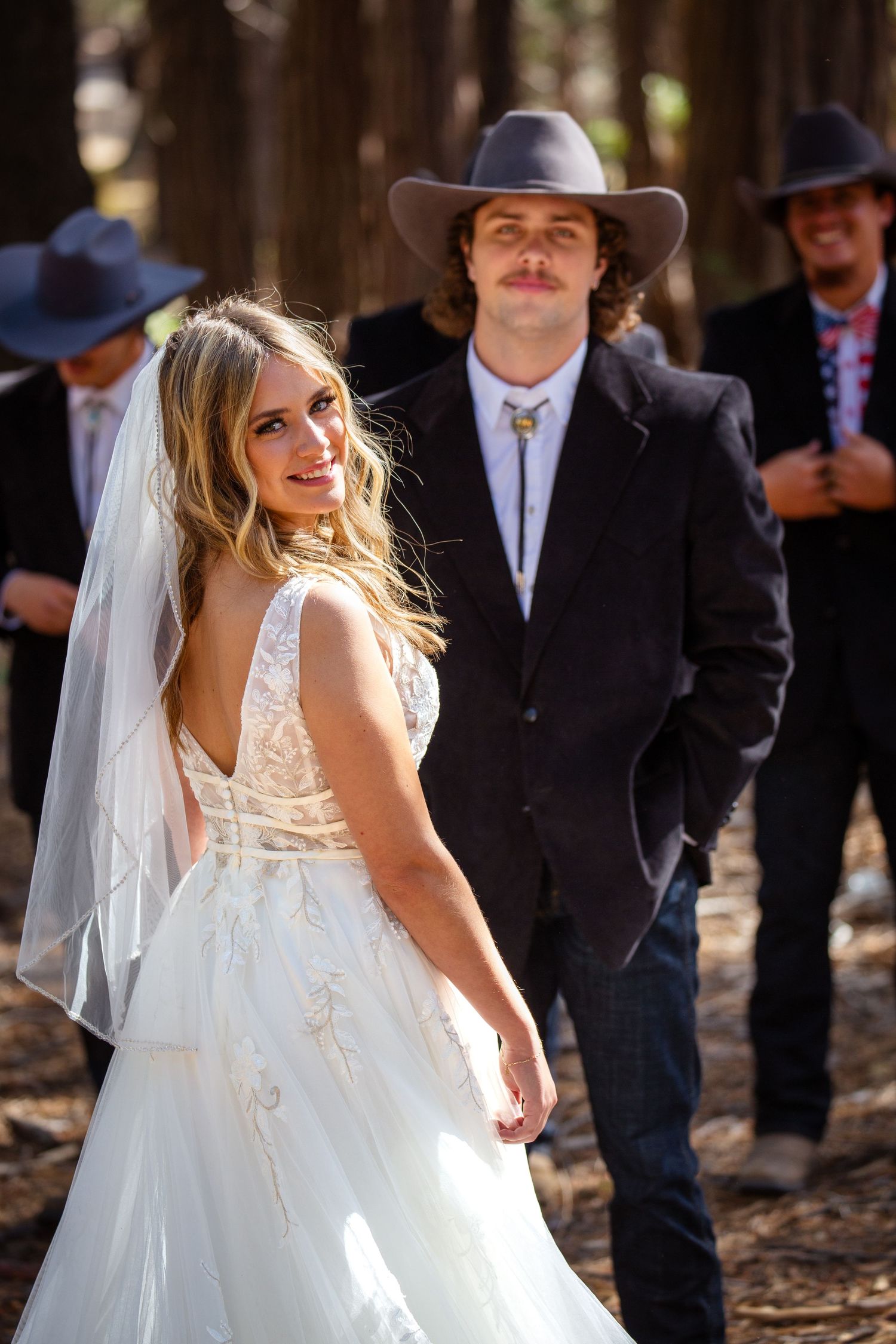 A rustic country wedding in the woods with a bride in a lace dress and groom in western attire with cowboy hats.