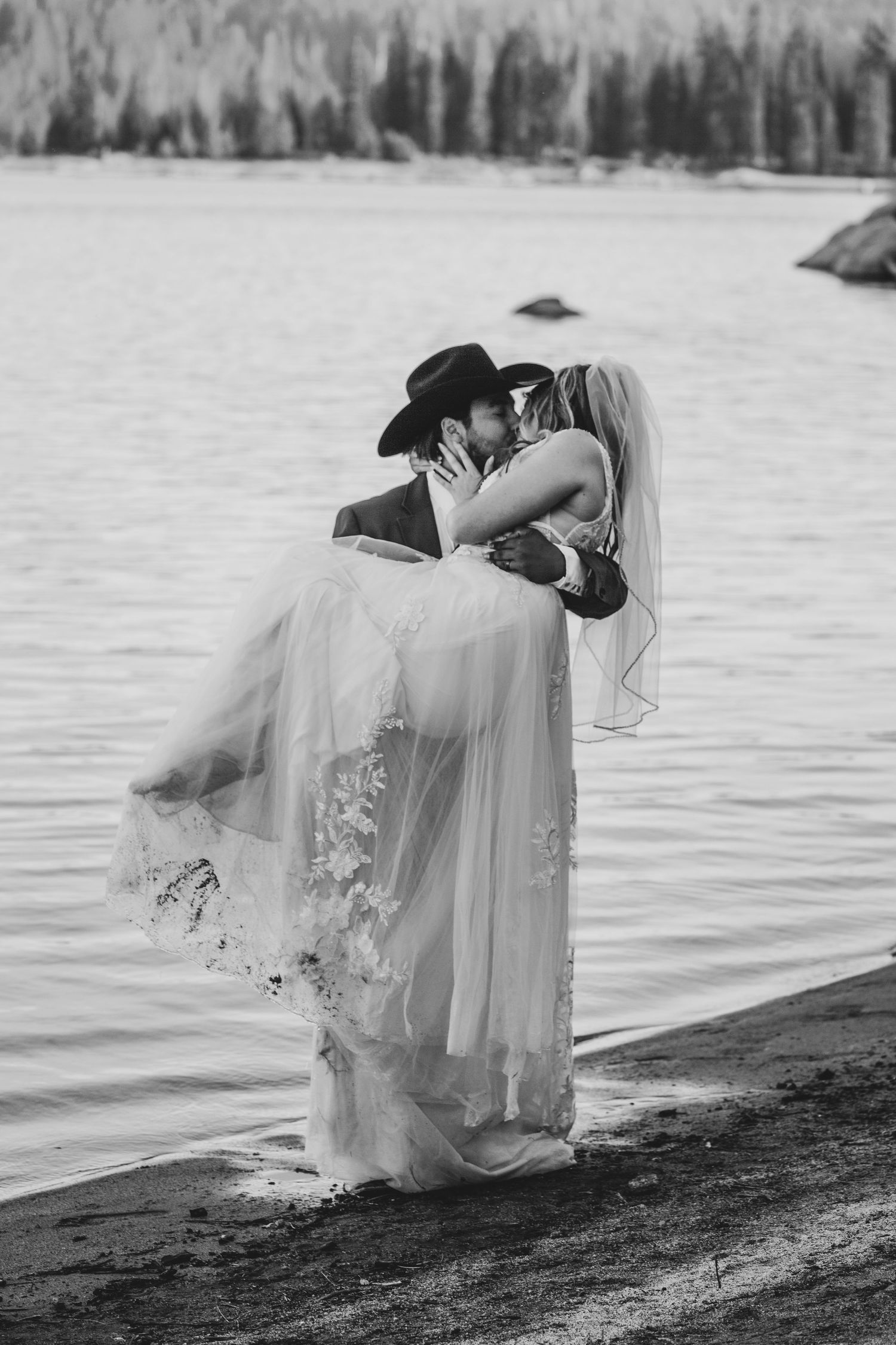 A romantic black and white wedding photo series showing a couple embracing by a lakeside with a flowing veil.