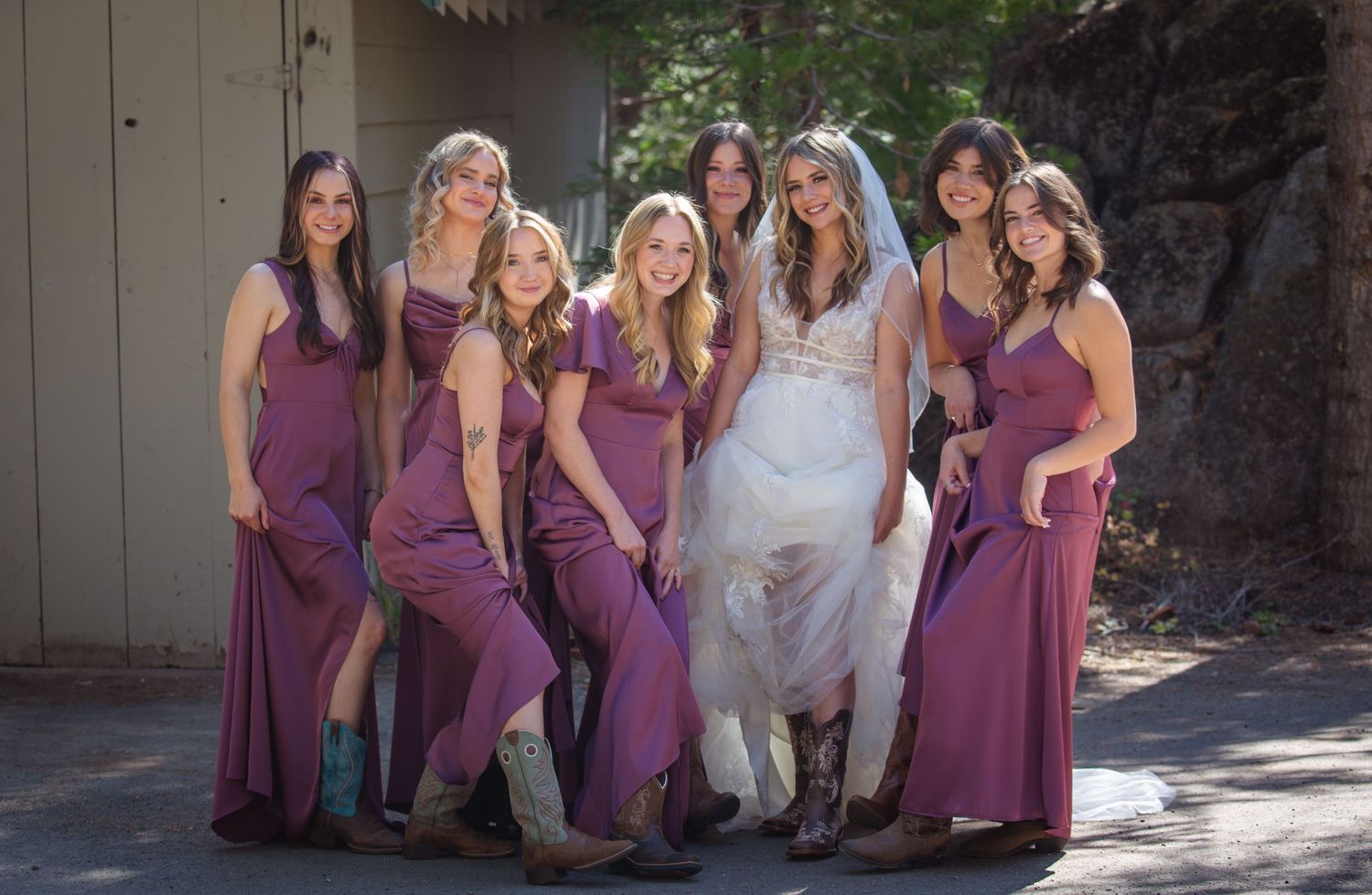Wedding party in matching burgundy dresses pose together with bride in white gown outdoors on sunny day.