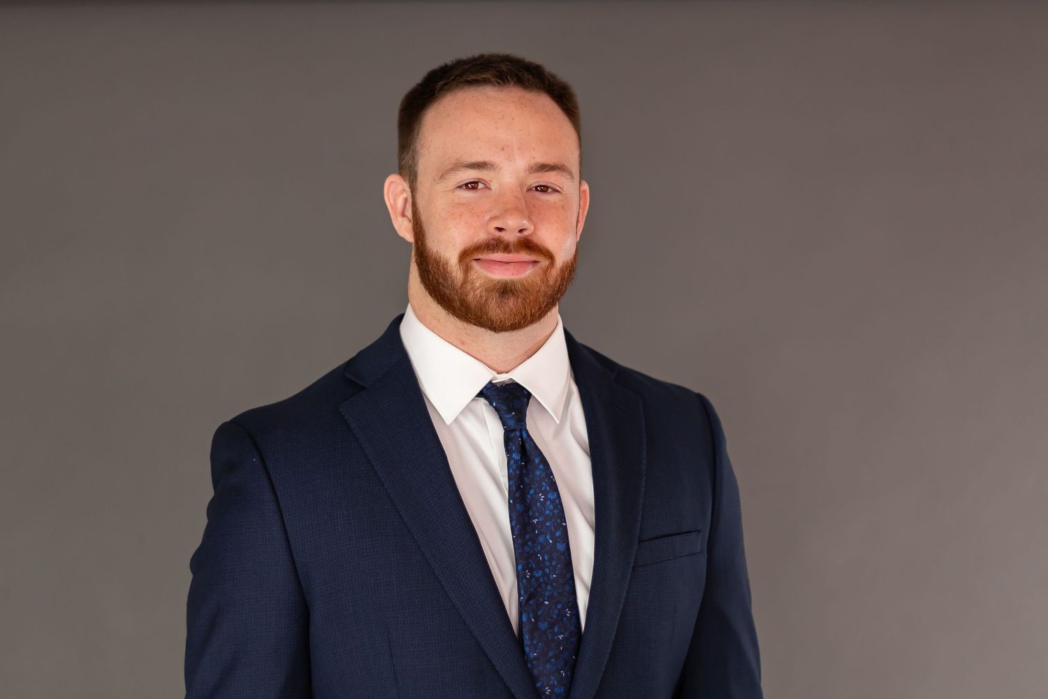 Professional headshot of someone wearing a navy suit and blue tie against a grey background.