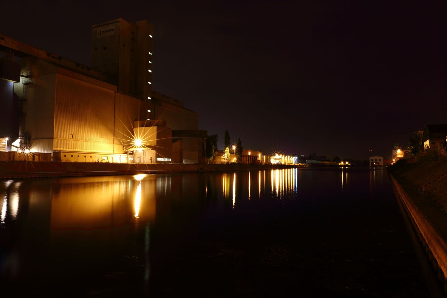 Industrial buildings reflect in calm water at night with glowing lights.