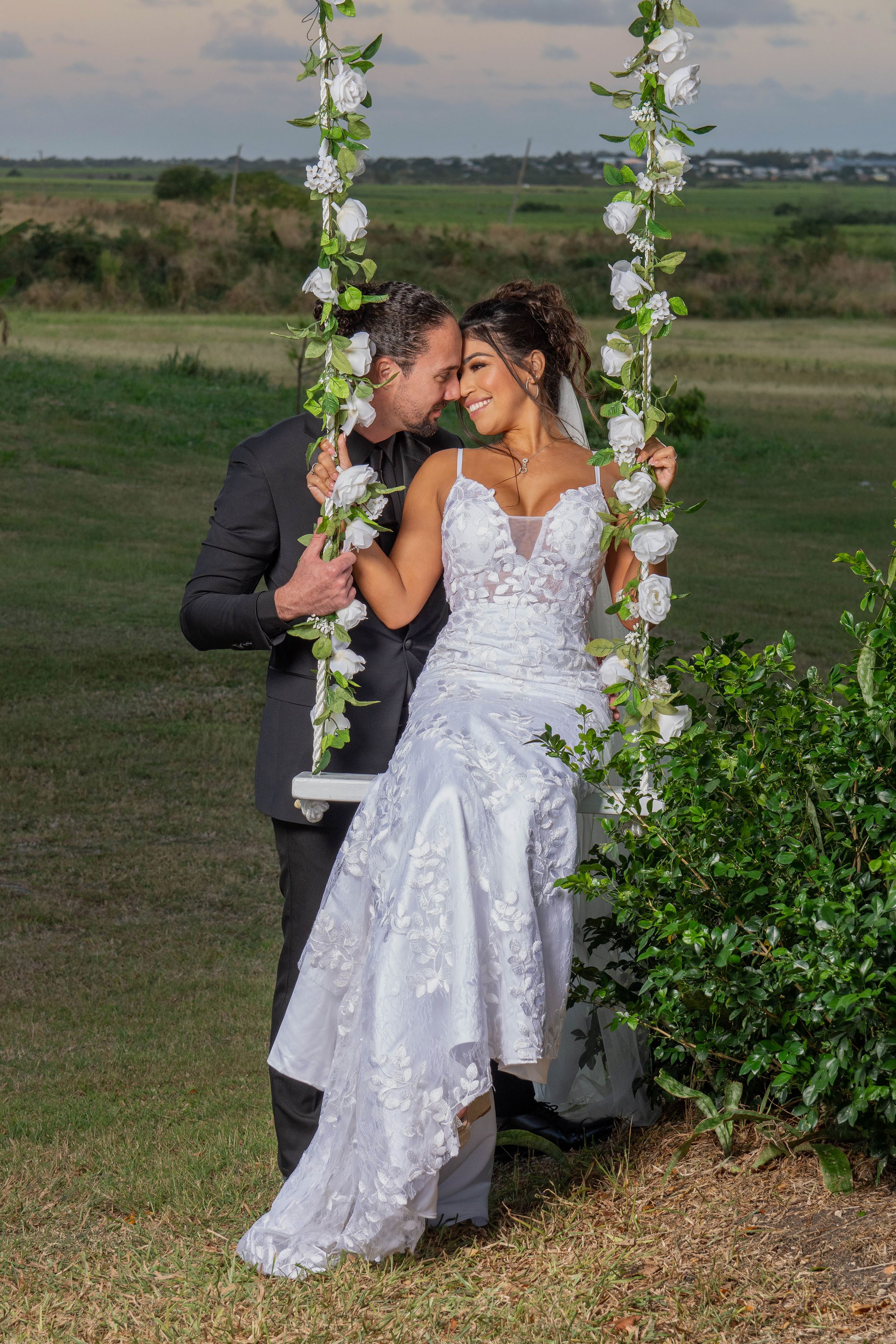 Bride in white lace gown sits on floral swing as groom in dark suit leans in close, with green fields behind.