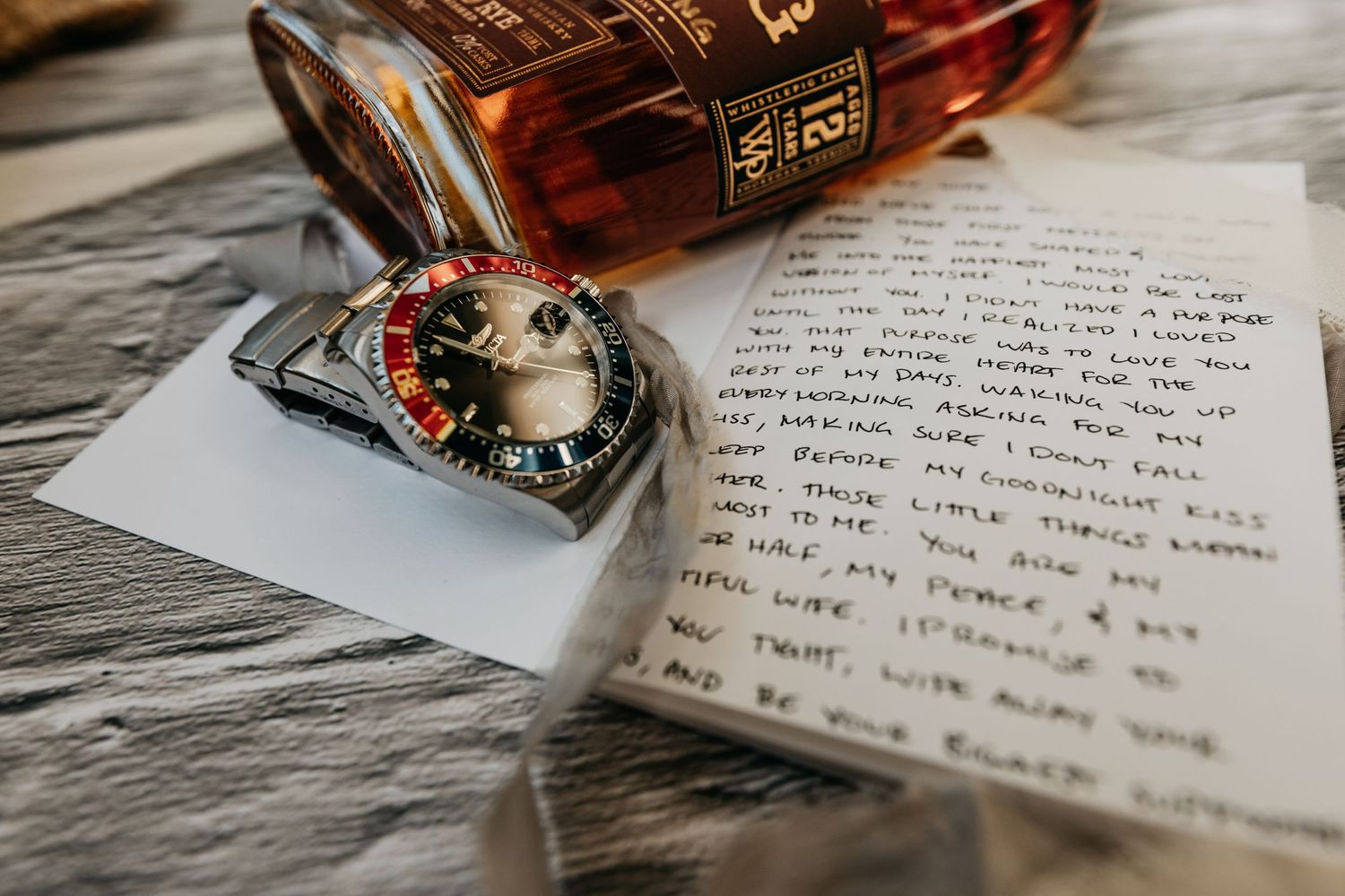 A handwritten wedding letter beside a luxury watch and bottle of bourbon on a rustic wooden surface.