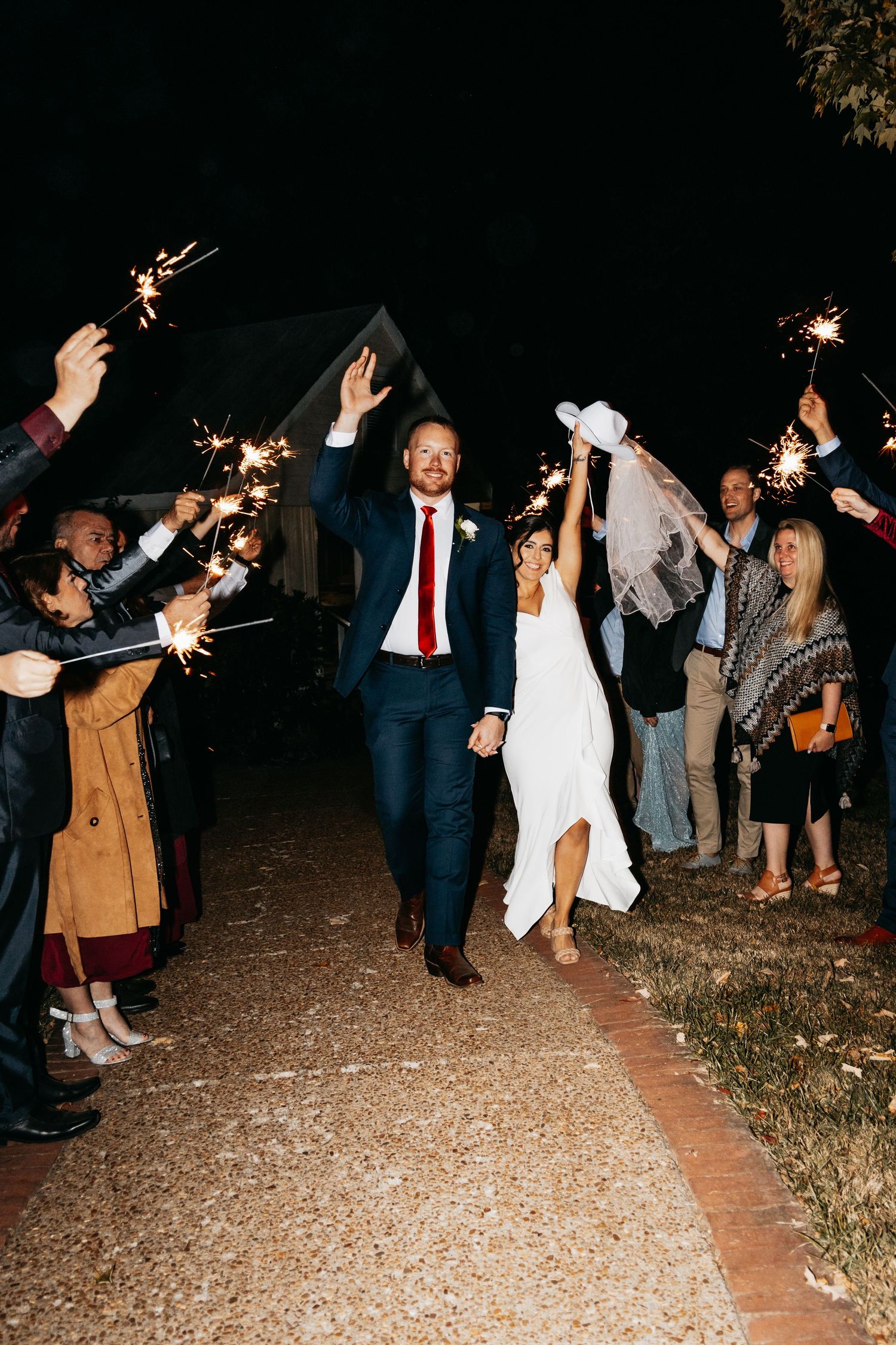 Joyful newlywed couple exits wedding ceremony through a sparkling sparkler send-off tunnel at night.