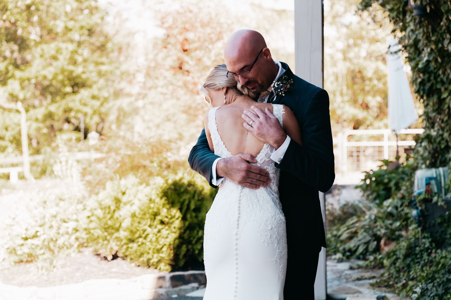 Bald groom embracing bride in a fitted lace wedding gown during first look.