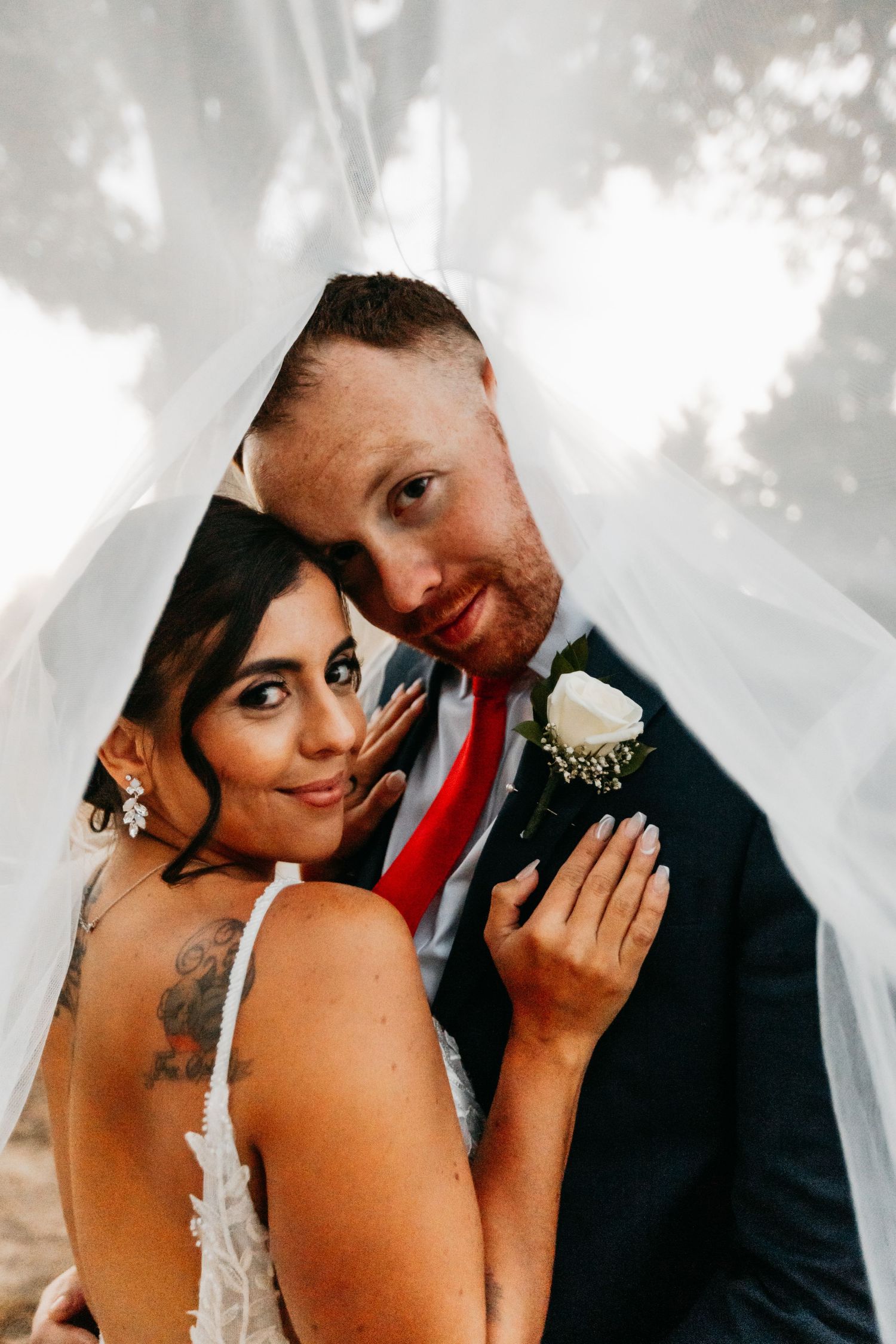 Bride and groom pose intimately together beneath a flowing white veil outdoors.