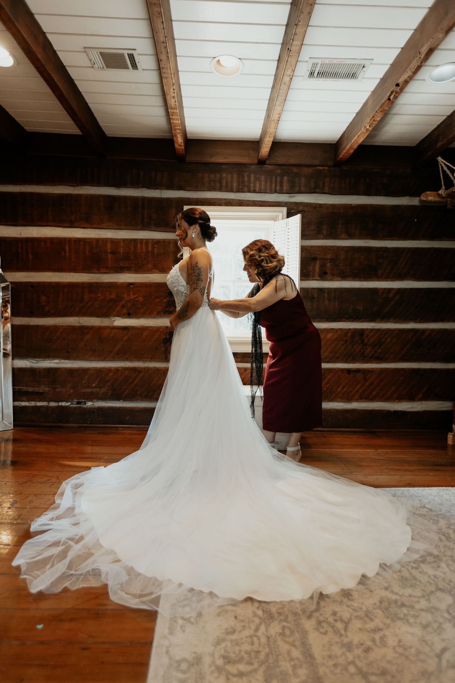 Mother helping bride fasten her elegant lace wedding gown in a rustic log cabin venue.