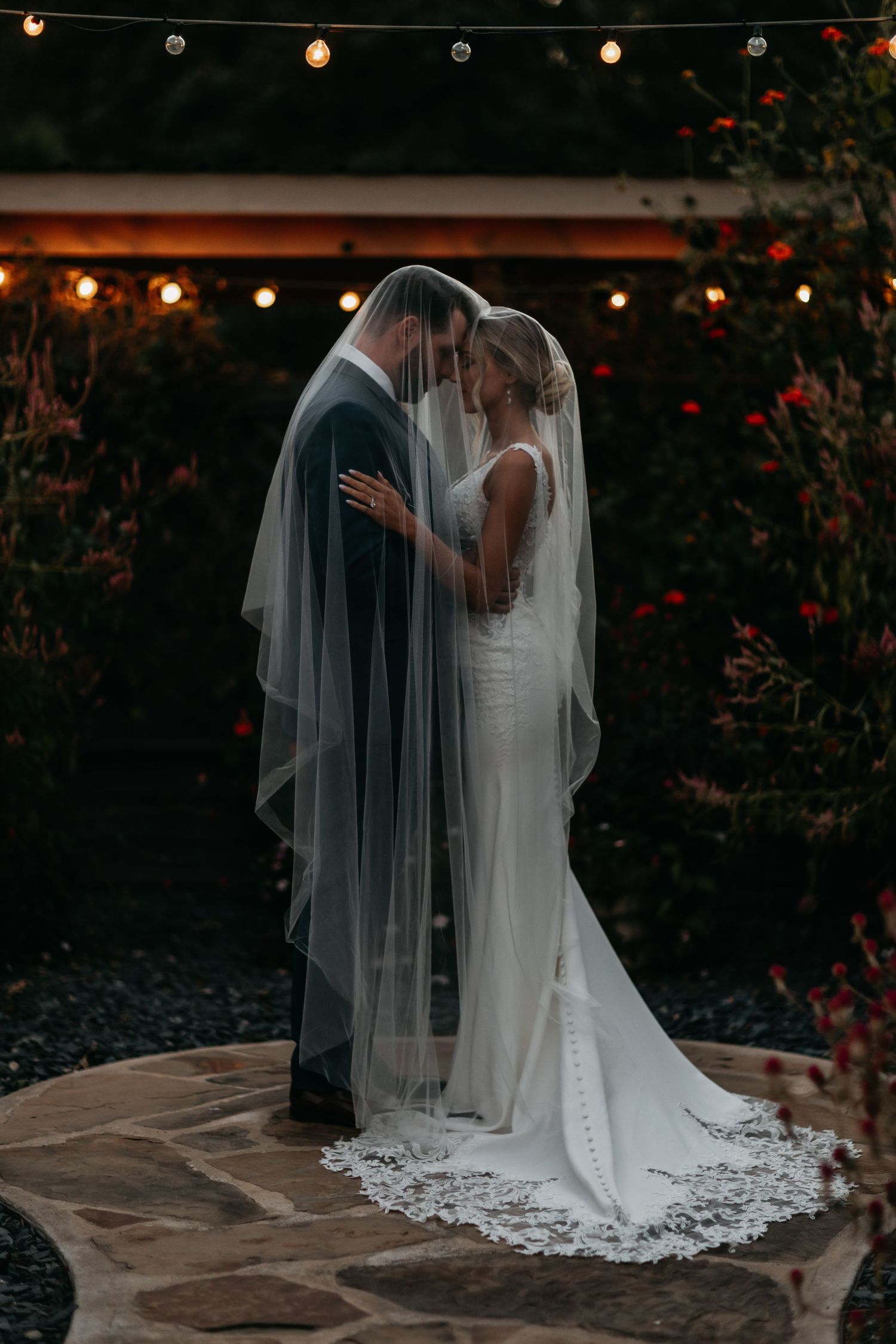 Bride and groom share an intimate moment under a flowing lace-trimmed veil surrounded by string lights at dusk.