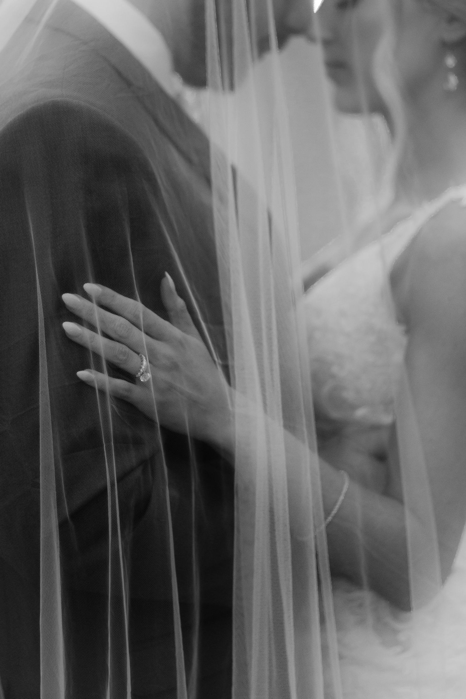 Black and white close-up of bride's hand with engagement ring resting on groom's suit beneath a sheer veil.