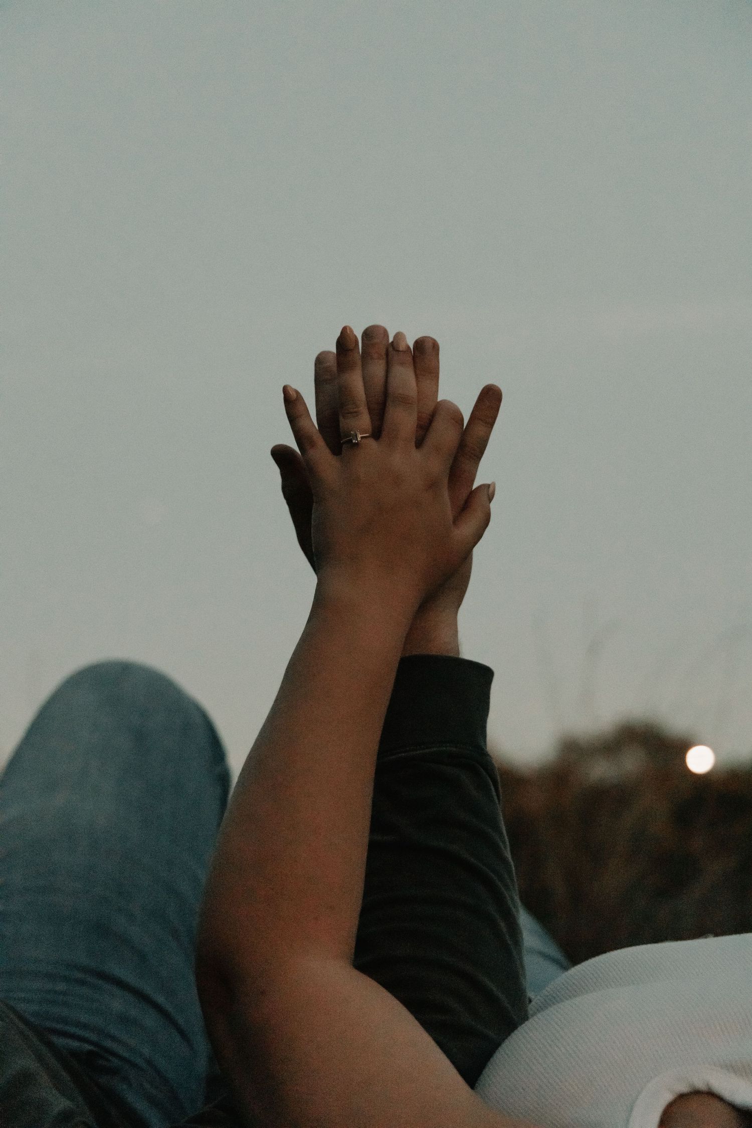 Two people holding hands with interlaced fingers raised against a soft evening sky.