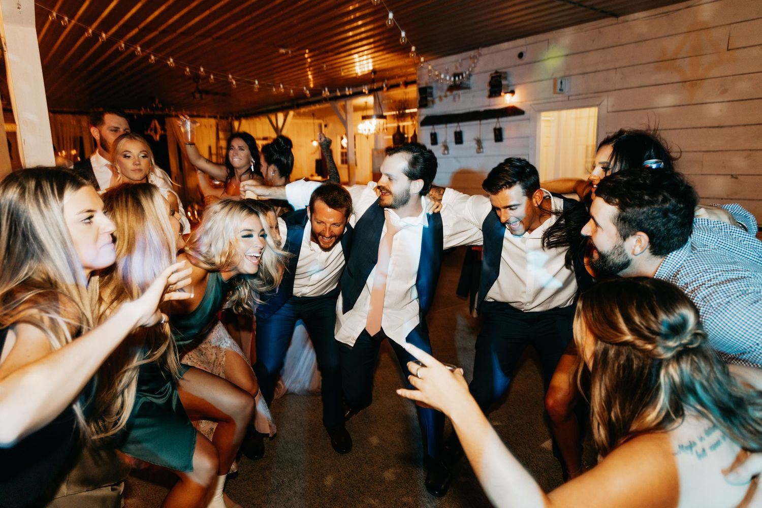 Guests dancing energetically at a lively wedding reception inside a rustic wooden venue.