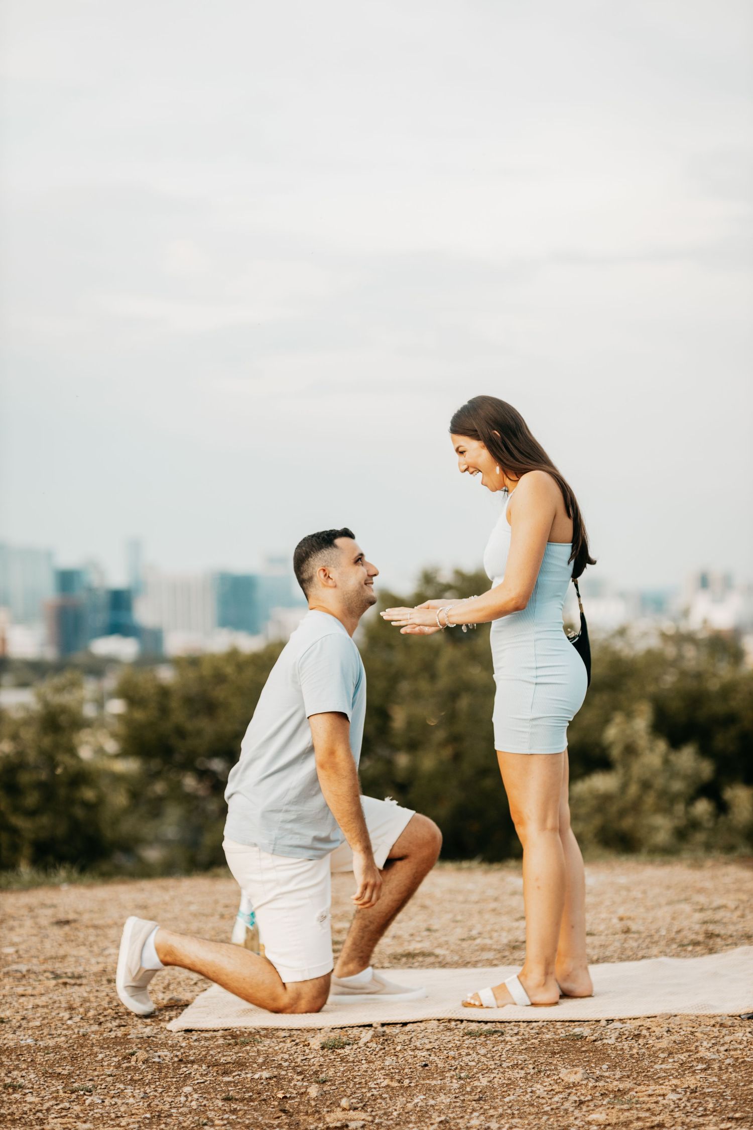 Man on one knee proposing to a smiling woman in a blue dress with a city skyline in the background.
