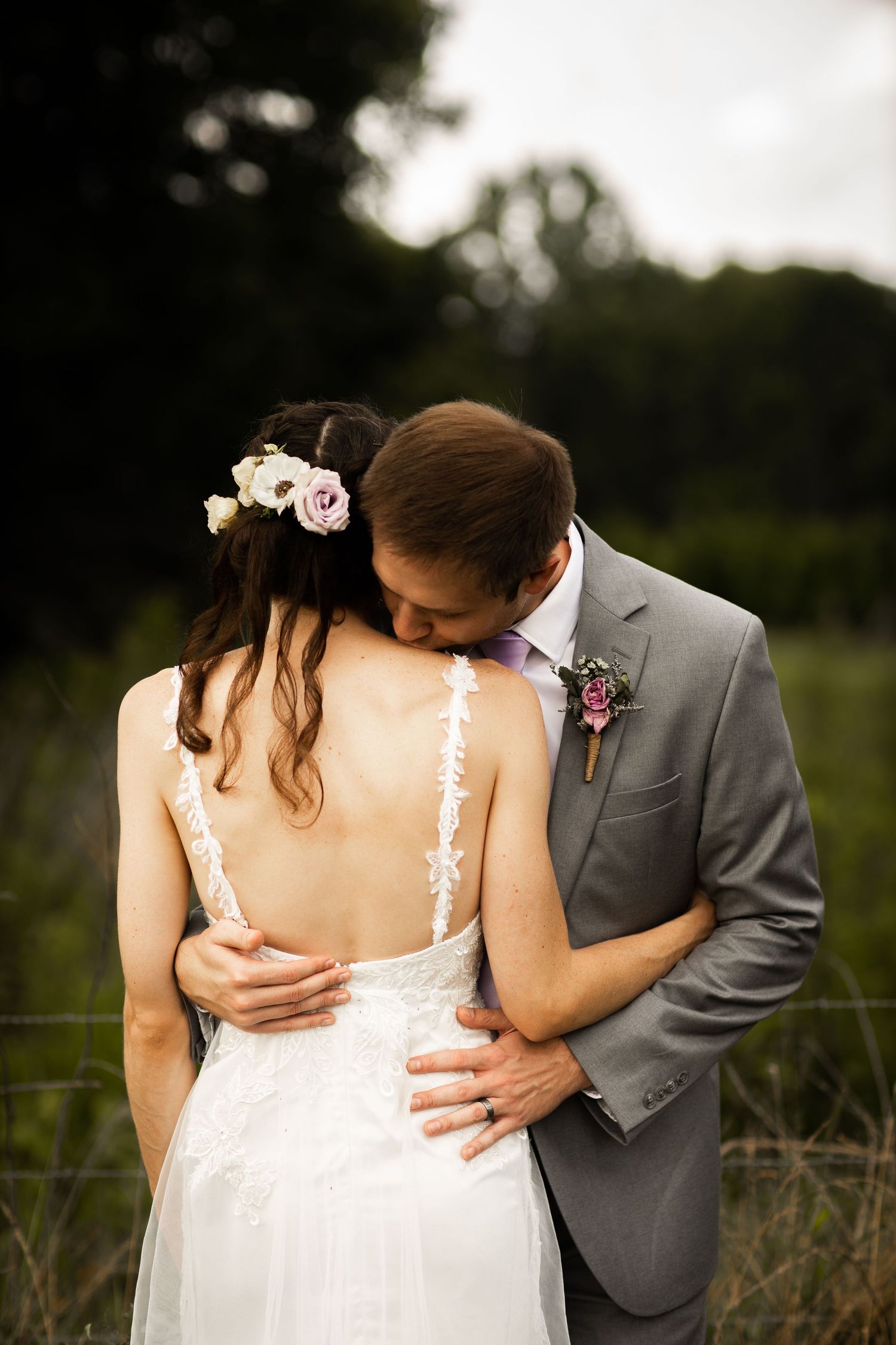 Groom in gray suit kisses bride wearing backless lace wedding dress with floral hair accessories.