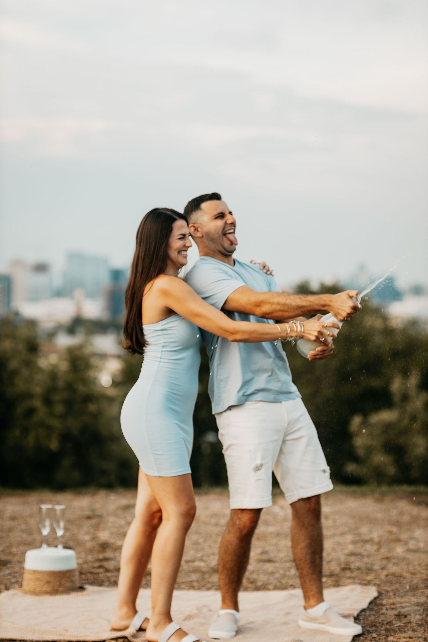 Joyful couple popping a champagne bottle together outdoors with a city skyline visible in the background.