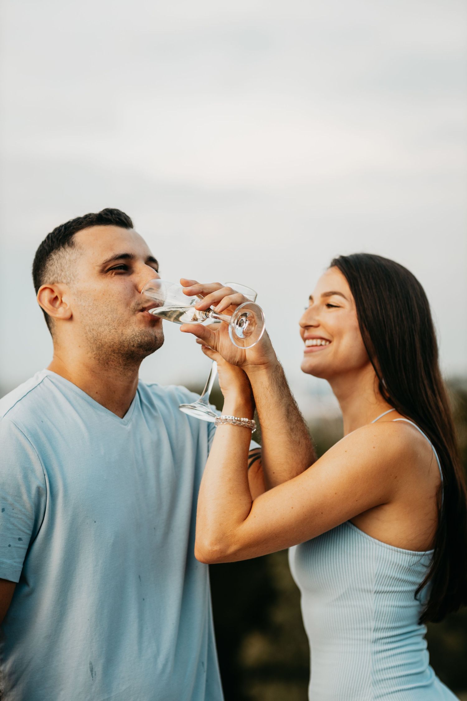 Smiling couple celebrating outdoors as woman helps man drink champagne from a stemmed glass.